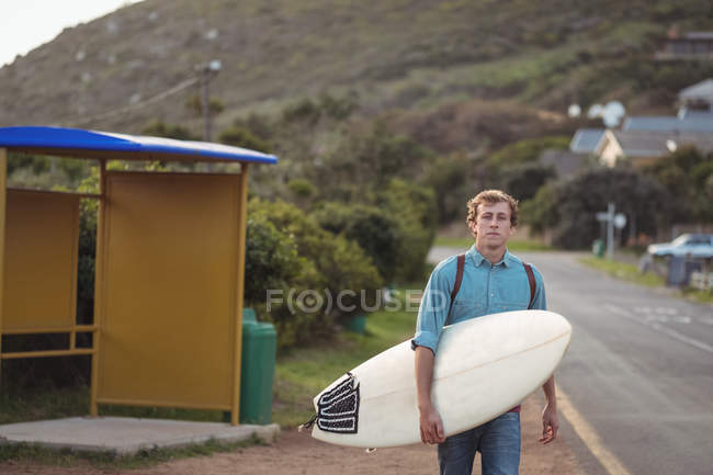 Portrait of a man carrying a surfboard walking along road — Stock Photo