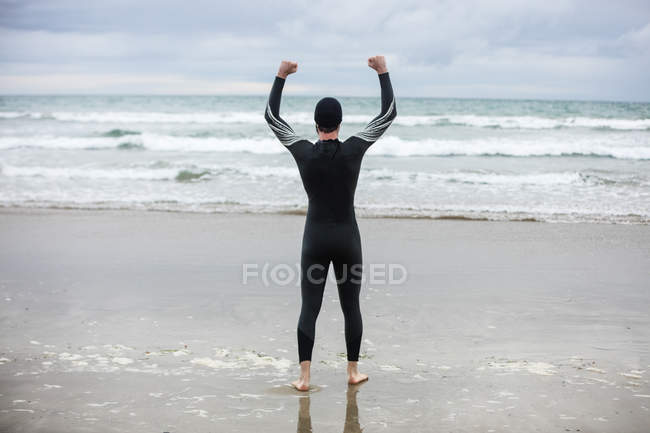 Rear view of athlete in wet suit standing with arms up on beach — Stock Photo