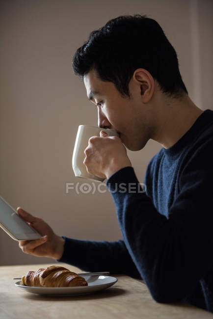 Homem usando tablet digital enquanto toma café da manhã em casa — Fotografia de Stock