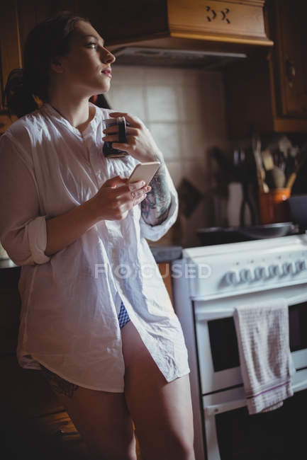 Продумана жінка використовує мобільний телефон, маючи каву на кухні вдома — стокове фото