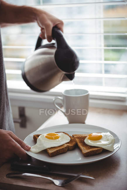 Мужчина держит тарелку с завтраком, наливая горячую воду в кружку на кухне. — стоковое фото