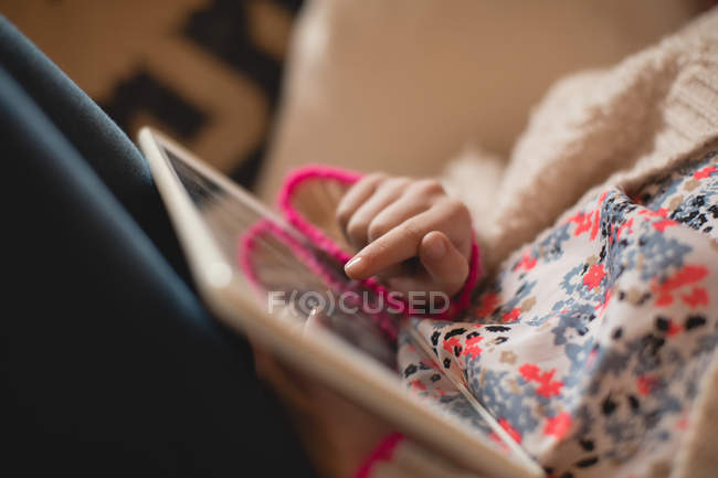 Sección media de la niña sentada en el sofá y el uso de la tableta digital en la sala de estar en casa - foto de stock