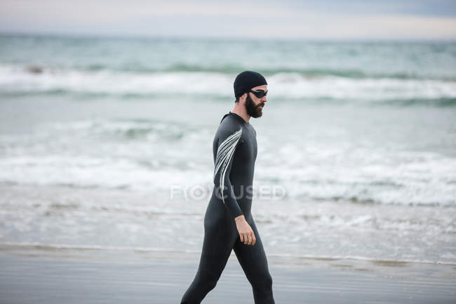 Guapo atleta en traje de neopreno caminando en la playa - foto de stock