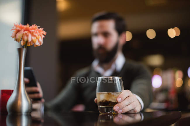 Hombre tomando un vaso de bebida mientras usa el teléfono móvil en el bar - foto de stock