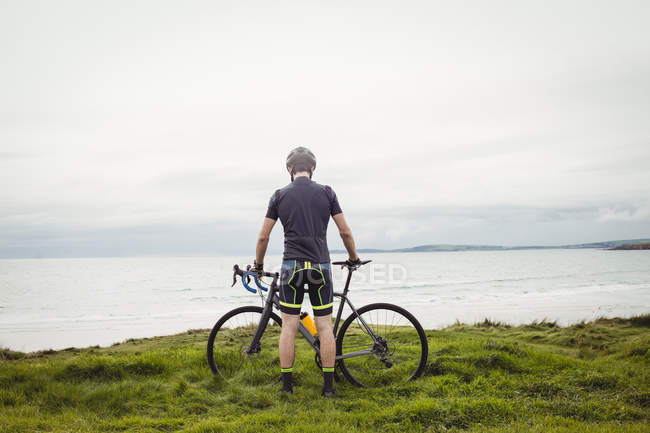 Atleta de pé com bicicleta na grama perto do mar — Fotografia de Stock