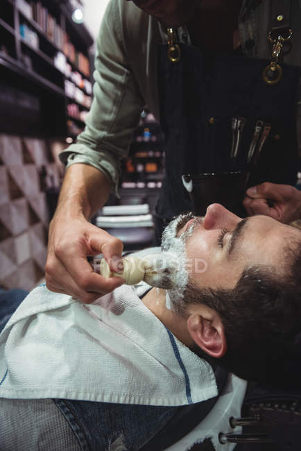 Парикмахер наносит крем на бороду клиента в парикмахерской — стоковое фото