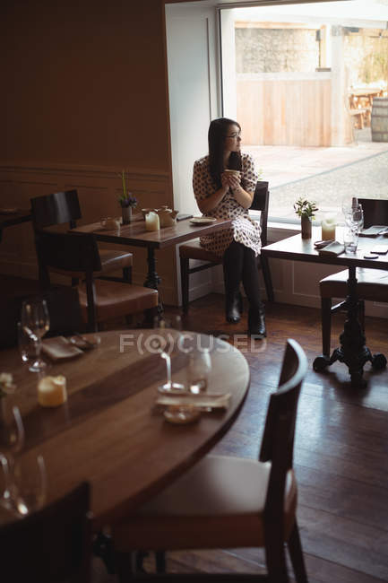 Mujer pensativa tomando una taza de café cerca de la ventana en el café - foto de stock