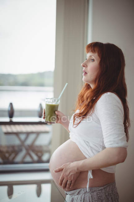 Pensativa mujer embarazada sosteniendo un vaso de jugo en casa - foto de stock