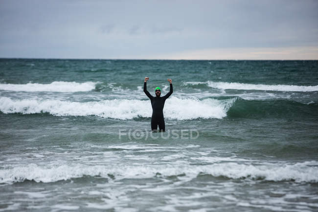 Atleta de terno molhado em pé no mar com as mãos levantadas — Fotografia de Stock
