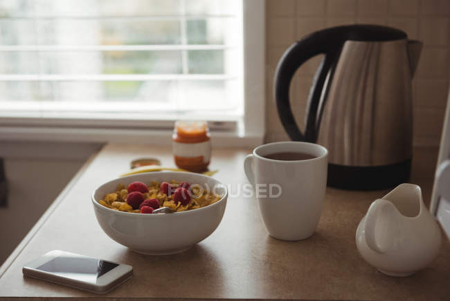 Cereales de desayuno con taza de café y teléfono móvil en la encimera de la cocina en casa - foto de stock