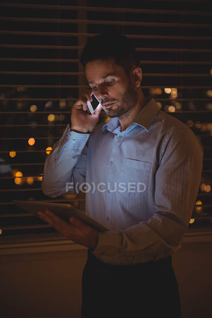 Homme parlant sur un téléphone portable tout en utilisant une tablette numérique près de la fenêtre la nuit — Photo de stock