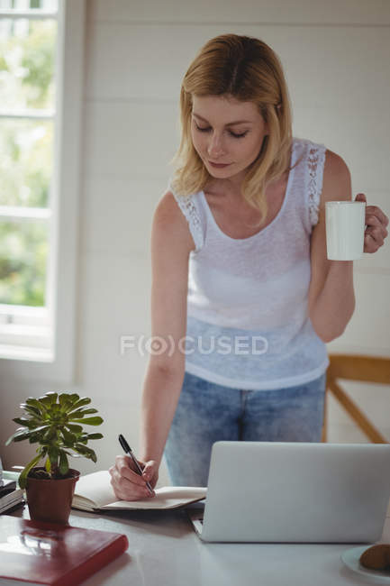 Hermosa mujer escribiendo en el diario mientras toma café en la sala de estar en casa - foto de stock