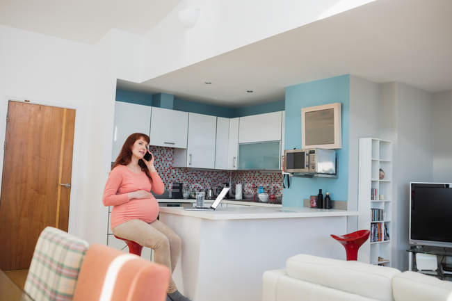 Pregnant woman talking on mobile phone in kitchen at home — Stock Photo