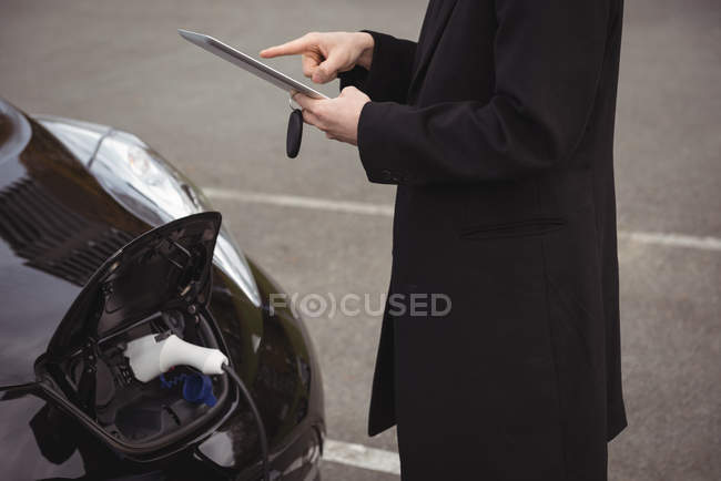 Hombre usando tableta digital mientras carga coche eléctrico en la estación de carga del vehículo eléctrico - foto de stock