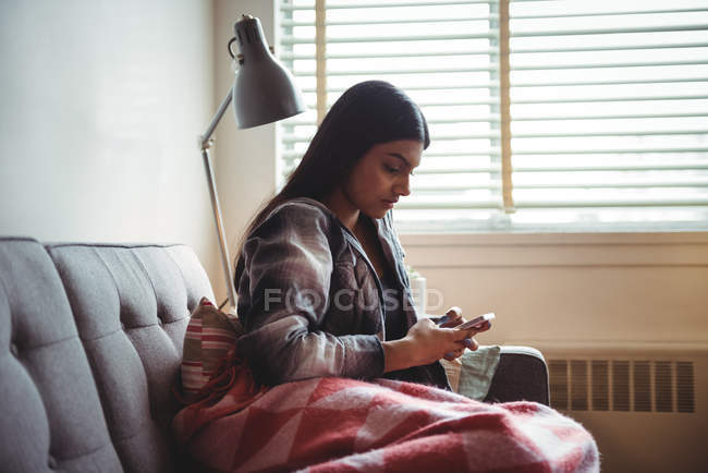 Mujer usando el teléfono móvil mientras se relaja en el sofá en la sala de estar en casa - foto de stock