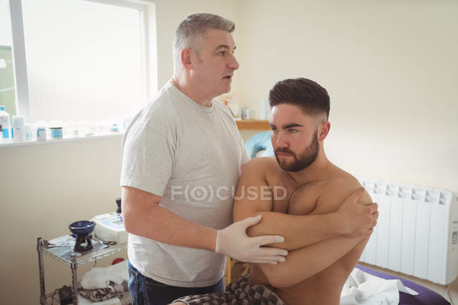 Fisioterapeuta maduro examinando hombro de paciente masculino en clínica - foto de stock