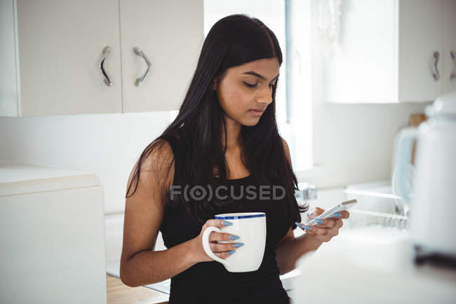 Mulher usando telefone celular enquanto segurando xícara de café na cozinha em casa — Fotografia de Stock