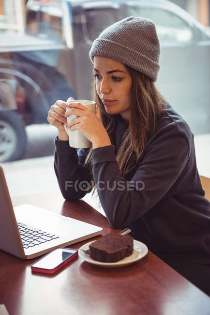 Mujer con ropa de invierno sosteniendo taza de café y mirando a la computadora portátil en el restaurante - foto de stock