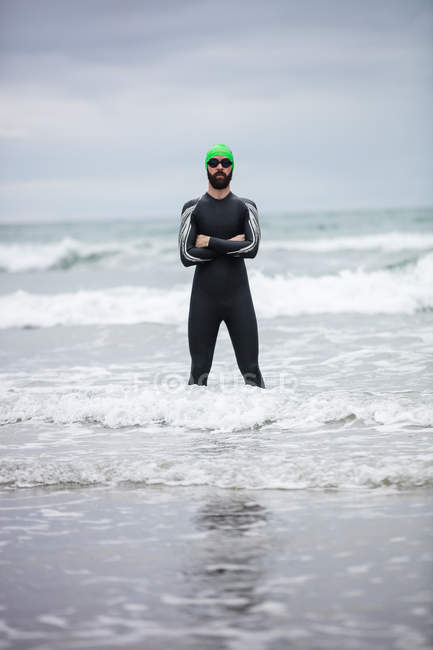 Спортсмен в мокром костюме стоит с руками, скрещенными в море на пляже — стоковое фото