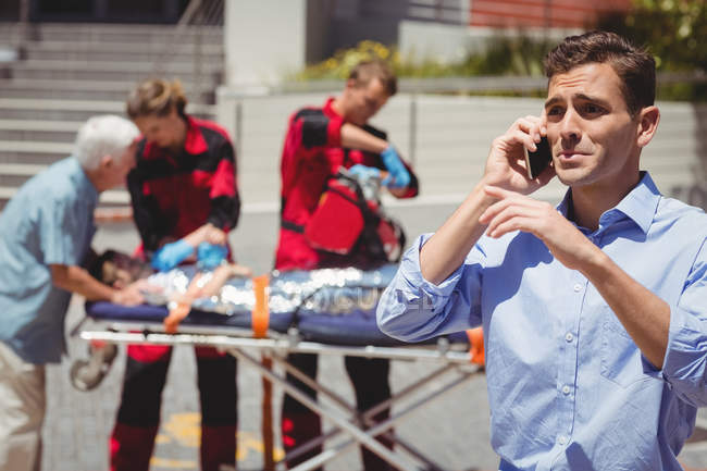 Homem falando no telefone celular e paramédicos examinando menino ferido na rua no fundo — Fotografia de Stock