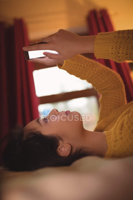 Femme couchée et utilisant un téléphone portable sur le lit dans la chambre à coucher à la maison — Photo de stock
