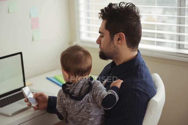 Vater hält sein Baby während der Handynutzung am Schreibtisch zu Hause — Stockfoto