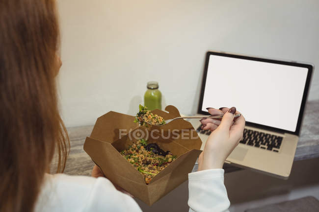 Vista trasera de la mujer comiendo ensalada mientras mira el portátil - foto de stock