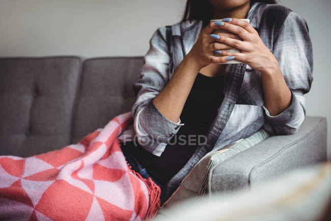 Seção média de mulher segurando xícara de café sentado no sofá na sala de estar em casa — Fotografia de Stock