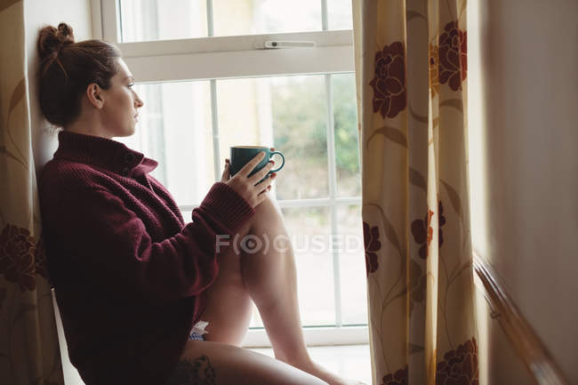 Mulher pensativo sentado na soleira da janela e segurando xícara de café em casa — Fotografia de Stock