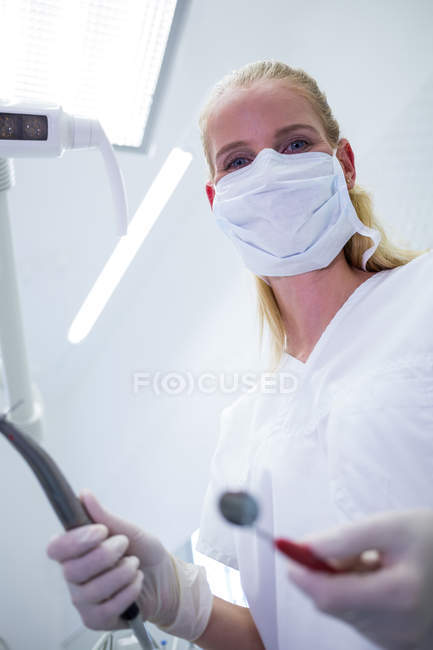 Dentista femenina con máscara quirúrgica que sostiene instrumentos dentales en la clínica - foto de stock