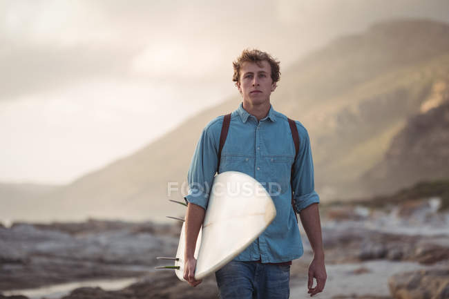 Portrait of a man carrying a surfboard walking by sea at dusk — Stock Photo