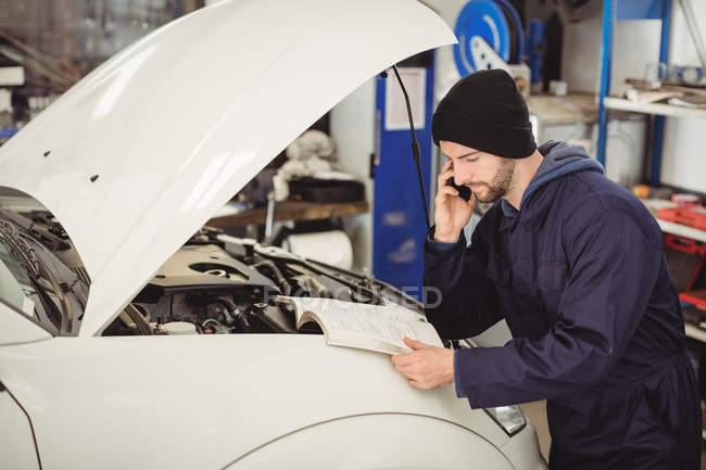 Mechanic reading instruction manual while talking on mobile phone at repair garage — Stock Photo