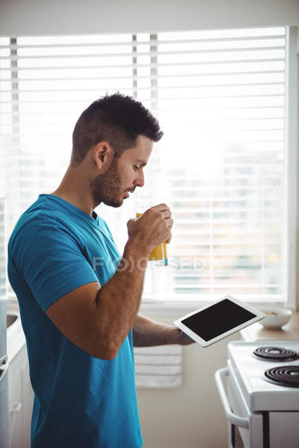 Mann benutzt sein digitales Tablet, während er zu Hause in der Küche ein Glas Saft trinkt — Stockfoto