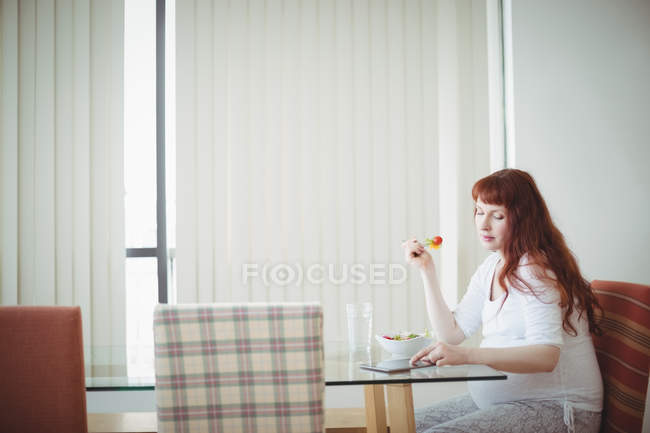 Mulher grávida usando comprimido digital enquanto come salada em casa — Fotografia de Stock
