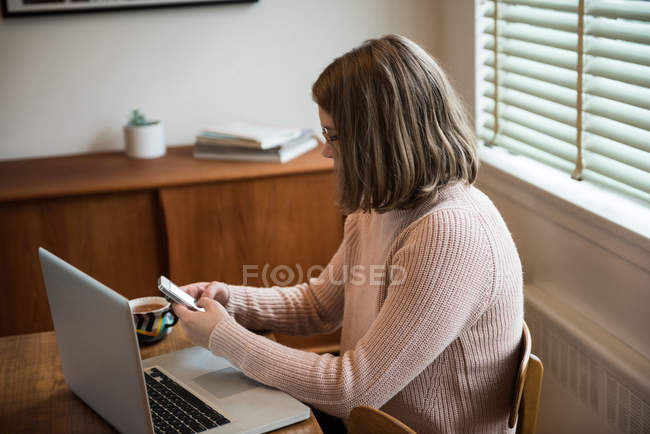 Mulher usando telefone celular enquanto trabalhava no laptop em casa — Fotografia de Stock