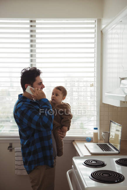 Отец разговаривает по мобильному телефону, держа ребенка на кухне — стоковое фото
