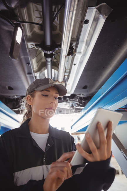 Женщина-механик с цифровым планшетом под автомобилем в ремонтном гараже — стоковое фото