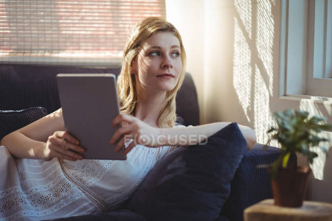 Thoughtful woman lying on sofa and holding digital table in living room at home — Stock Photo