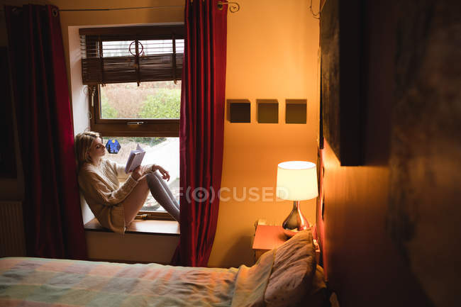 Женщина сидит на окне и читает книгу дома — стоковое фото