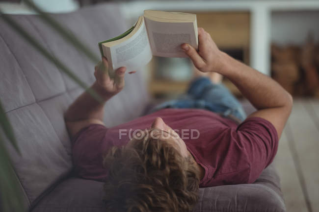 Человек лежит на диване и читает книгу дома — стоковое фото