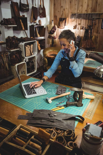 Женщина пользуется ноутбуком во время разговора по мобильному телефону в мастерской — стоковое фото