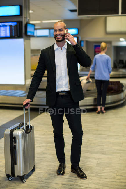 Empresario con equipaje hablando por teléfono móvil en zona de espera en terminal aeroportuaria - foto de stock