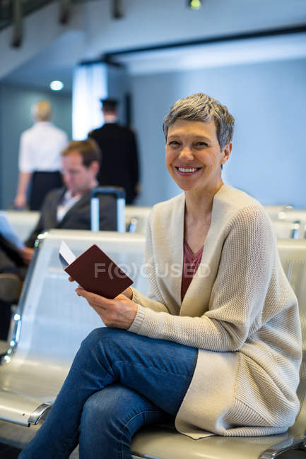 Retrato de mujer sonriente con pasaporte sentado en la sala de espera en la terminal del aeropuerto - foto de stock