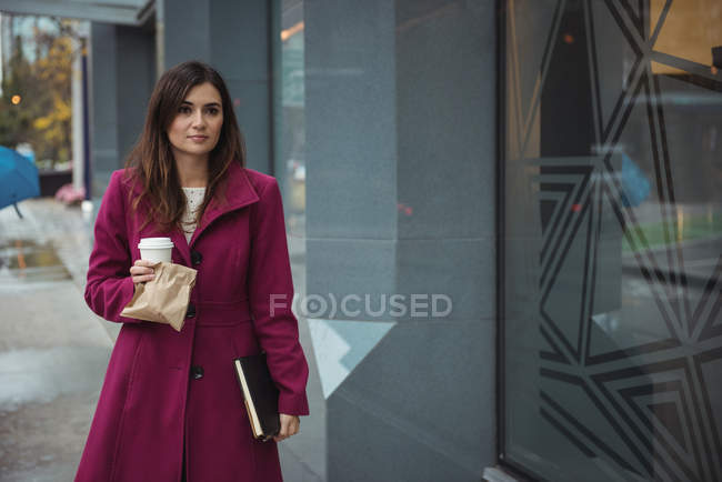 Empresária segurando copo de café descartável, pacote e diário andando na calçada — Fotografia de Stock