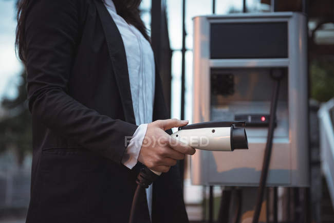 Средняя секция женщины, держащей зарядное устройство для автомобиля на электростанции зарядки — стоковое фото