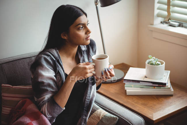Donna che tiene la tazza di caffè guardando attraverso la finestra a casa — Foto stock