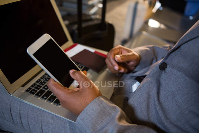 Sección media del hombre con el ordenador portátil usando el teléfono mientras está sentado en la terminal del aeropuerto - foto de stock
