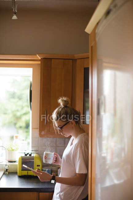 Femme utilisant une tablette numérique tout en prenant un café dans la cuisine à la maison — Photo de stock