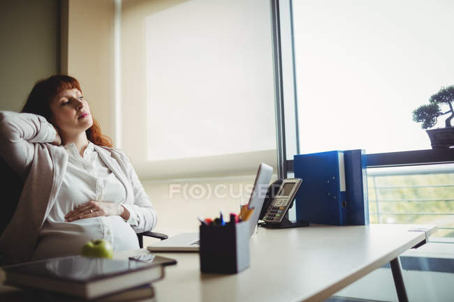 Вагітна бізнес-леді тримає біль назад, сидячи на стільці в офісі — стокове фото