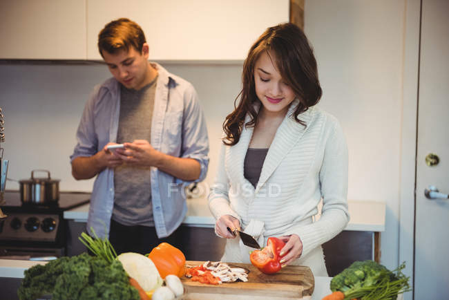 Женщина режет овощи и мужчина с помощью мобильного телефона на кухне дома — стоковое фото